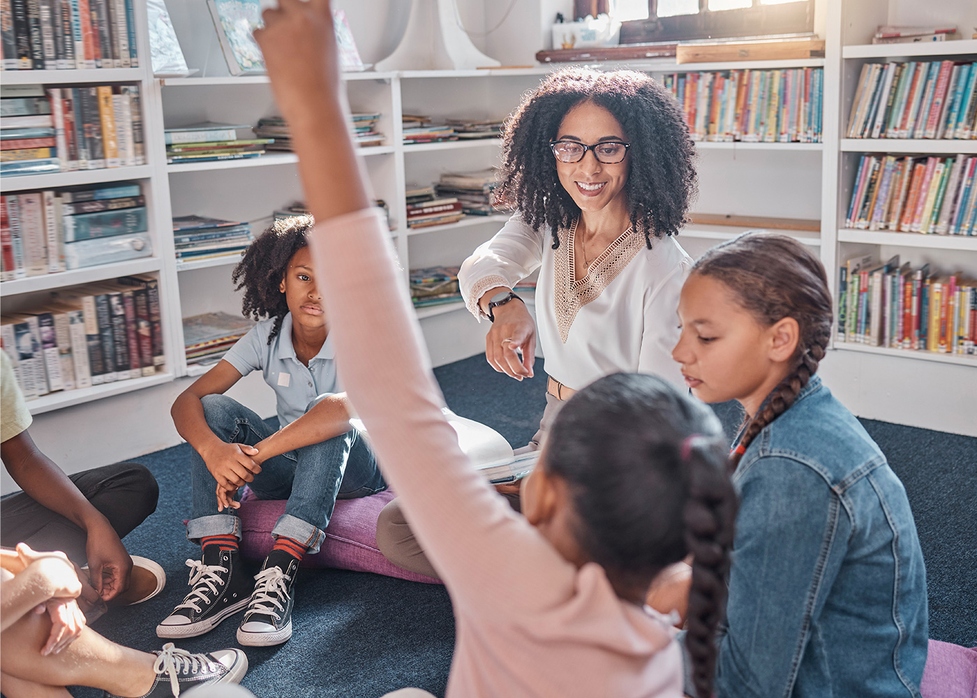 children in classroom sitting near bookshelves and interacting with teacher